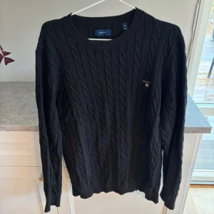 Svart kabelstickad tröja från GANT - Säljer en stilren svart kabelstickad tröja från GANT. Tröjan har en klassisk rund halsringning och långa ärmar. Perfekt för kyligare dagar och ger en tidlös look. Passar bra till både jeans och chinos.