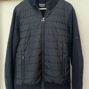 Barbour Cardigan marinblå - Snygg marinblå cardigan från Barbour med dragkedja och ribbad krage i bra skick. Tröjan har långa ärmar och två praktiska fickor med dragkedja framtill. Perfekt för en stilren look. Nypris 2200kr.