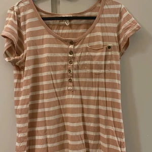 Randig kortärmad topp - Säljer en randig kortärmad topp i beige och vit med knappar framtill och en bröstficka. Perfekt för en avslappnad stil.