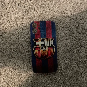 Mobilskal med FC barcelona loggan på - Snyggt iPhone 12 min fodral med FC Barcelonas logga i rött och blått. Skalet har en stilren design med vertikala ränder och klubbens emblem i mitten. Perfekt för fotbollsfans som vill visa sitt stöd för laget. Bara lite sönder på kanten