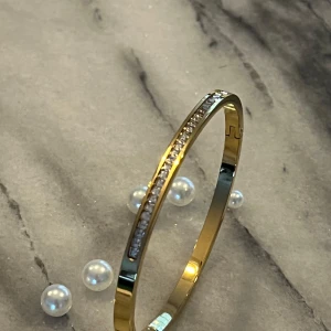 Guldarmband i rostfritt stål - Elegant guldarmband med en rad av gnistrande stenar längs ovansidan. Perfekt för att ge en touch av glamour till din outfit. Armbandet har en slät och glänsande yta som fångar ljuset vackert. Nytt och oanvänt! 