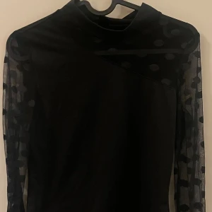 Svart topp med prickiga meshärmar - Snygg svart topp med unika prickiga meshärmar och asymmetrisk design. Perfekt för en stilren look med en twist. Toppen har en elegant passform och är tillverkad i ett mjukt material.
