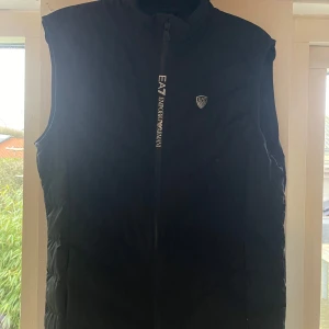 Svart väst från Emporio Armani - Snygg svart dunväst från Emporio Armani med dragkedja framtill och EA7-logga på bröstet. Perfekt för kyligare dagar med sin quiltade design som ger extra värme. Stilren och enkel att matcha med olika outfits.