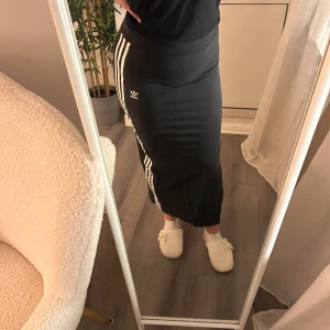 Långkjol från Adidas - Snygg svart långkjol från Adidas med de klassiska tre vita ränderna längs sidorna. Kjolen har en slits på sidan för extra rörelsefrihet och en tight passform som framhäver figuren. Perfekt för en sportig och stilren look.