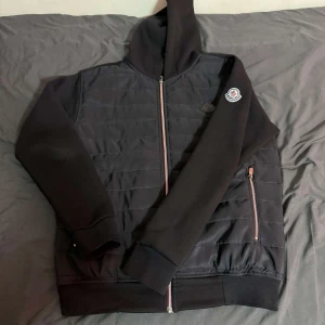 Svart cardigan med dragkedja från Moncler - Snygg svart cardigan från Moncler med dragkedja och huva. Den har ett quiltat mönster på framsidan och en broderad logga på ärmen. Perfekt för en stilren look.