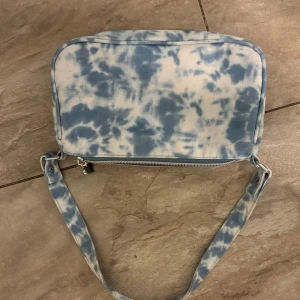 Blå och vit axelväska med batikmönster - Snygg axelväska i blått och vitt med coolt batikmönster. Väskan har en dragkedja med ett hjärtformat hänge och ett justerbart axelband. Perfekt för att ge din outfit en unik touch!