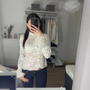 Vit spetsblus - Säljer en elegant vit spetsblus med långa ärmar och hög hals. Blusen har ett vackert blommönster och är perfekt för en stilren look. Passar bra till både jeans och kjol.