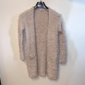 Beige kofta från Jacqueline de Yong - Mysig beige kofta från Jacqueline de Yong i en fluffig och mjuk design. Koftan har långa ärmar och två praktiska fickor framtill. Perfekt för att hålla sig varm och stilren under kyligare dagar.