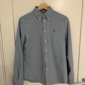 Blå skjorta från Polo Ralph Lauren - Snygg blå skjorta från Polo Ralph Lauren med klassisk krage och broderad logga på bröstet. Skjortan har långa ärmar och knappar framtill. Perfekt för en stilren look.