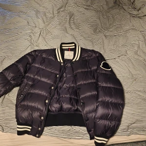 Svart pufferjacka från Moncler - Säljer en snygg svart pufferjacka från Moncler med vita detaljer. Jackan har långa ärmar och en ribbad krage samt muddar. Den är försedd med knappar och dragkedja framtill. Perfekt för kyligare dagar!