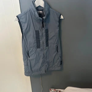 Blå väst från Stone Island - Snygg blå  väst från Stone Island med dragkedja och hög krage. Västen har praktiska fickor med dragkedja och den ikoniska loggan på sidan. Perfekt för lager-på-lager-stil. Storlek junior men sitter som S såklart äkta 