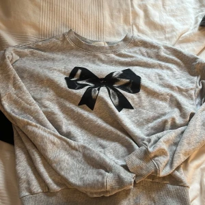 Grå tröja med svart rosett från H&M - Säljer en grå tröja från H&M med en stor svart rosett på framsidan. Tröjan har långa ärmar och en rund halsringning. Perfekt för en avslappnad stil.
