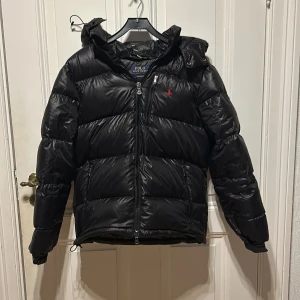 Svart dunjacka från Polo Ralph Lauren - Säljer en svart dunjacka från Polo Ralph Lauren med huva och dragkedja. Jackan har en broderad röd logga på bröstet och är perfekt för kalla dagar. Den är långärmad och har en stilren design. Säljer pga. Förstor, använd fåtal gånger pris är diskuterbart