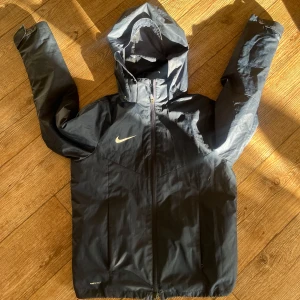Svart vindjacka från Nike - Säljer en svart vindjacka från Nike med huva och dragkedja. Jackan har en diskret Nike-logga på bröstet och är tillverkad i ett lätt syntetmaterial. Perfekt för blåsiga dagar.