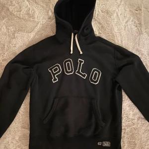 Svart hoodie från Polo Ralph Lauren - Säljer en riktigt snygg svart hoodie från Polo Ralph Lauren. Den har en klassisk känguruficka och justerbar huva med vita snören. Perfekt för en avslappnad stil.