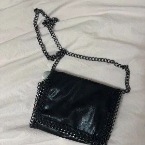Svart axelväska med kedjedetaljer - Snygg svart axelväska med kedjedetaljer runt kanterna och en kedja som axelrem. Väskan har en dragkedja inuti för extra säkerhet. Perfekt för att ge en edgy touch till din outfit. Knappen har gått sönder men dragkedjan funkar perfekt och inget kommer trilla ut. 