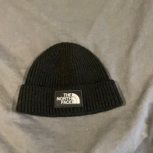 Svart ribbad mössa från The North Face - Snygg svart ribbad mössa från The North Face. Perfekt för kyliga dagar med sin stilrena design och varma material. Märkeslogga framtill ger en cool touch. Passar de flesta tack vare one size-storleken.