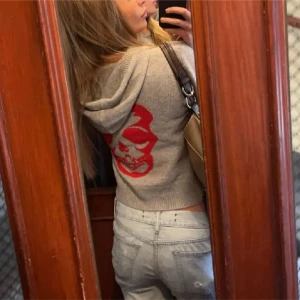 Grå hoodie från Zadig & Voltaire - Säljer en grå stickad hoodie från Zadig & Voltaire med en stor röd dödskalle på ryggen och en liten röd ZV-logga på framsidan. Tröjan har en praktisk magficka och en bekväm huva. Perfekt för en cool och avslappnad stil. Skulle säga att den är storlek XS/XXS