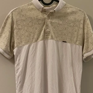 Beige och vit pikétröja från Bentner - Säljer en stilren beige och vit pikétröja från Bentner. Tröjan har korta ärmar och en klassisk krage med knappar. Perfekt för en avslappnad men ändå snygg look.