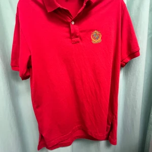 Röd pikétröja från Ivanhoe - Säljer en snygg röd pikétröja från Ivanhoe med broderad detalj på bröstet. Tröjan har korta ärmar och en klassisk krage med knappar. Perfekt för en stilren look.        