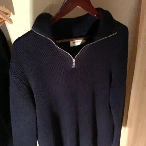 Mörkblå stickad halvzip tröja från East West - Säljer en snygg mörkblå stickad tröja från East West. Tröjan har en dragkedja vid halsen och långa ärmar, perfekt för kyligare dagar. Den är både stilren och bekväm, idealisk för en avslappnad look.