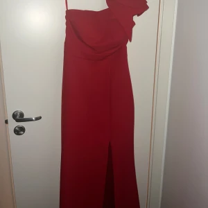 Röd axelbandslös klänning med volangdetalj - Elegant röd klänning med en axelbandslös design och volangdetalj vid axeln. Klänningen har en lång siluett med en slits framtill, perfekt för en festlig kväll. Den faller fint ner till marken och har en underbar passform. Klänningen har endast använts vid ett tillfälle och är i nyskick. Tjejen på bilden är 165 cm och hade ungefär 4 cm klack. Klänningen är i storlek 34/36
