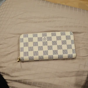 Plånbok - Snygg plånbok med ett klassiskt rutigt mönster i beige och grått. Plånboken har en praktisk dragkedja och flera fack för kort och kontanter. Perfekt för att hålla ordning på dina värdesaker.