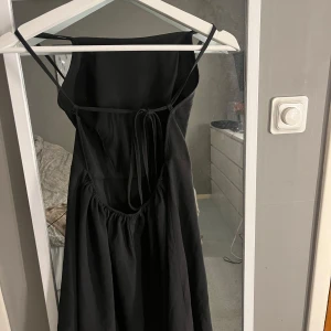 Svart axelbandslös klänning - Elegant svart klänning med smala axelband och en stilren design. Klänningen har en v-ringad fram och knytning i ryggen för en justerbar passform. Perfekt för en stilfull kväll.