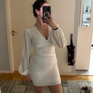 Vit kortklänning med v-ringning - Snygg vit kortklänning med v-ringning och långa ärmar. Klänningen har en figurnära passform och är perfekt för en stilren look. Den är enkel men elegant och kan stylas på många sätt.