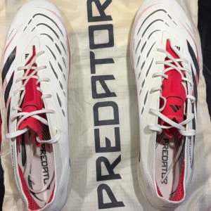 Adidas Predator fotbollsskor - Snygga Adidas Predator fotbollsskor i vitt med röda och svarta detaljer till konstgräs. Skorna är endast använda 2 pass och är i toppskick. Storlek 46. Nypris ligger på 3000kr