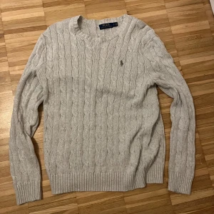 Grå kabelstickad tröja från Ralph Lauren - Säljer en stilren grå kabelstickad tröja från Ralph Lauren. Tröjan har en klassisk rund halsringning och långa ärmar. Perfekt för kyligare dagar med sin mjuka och bekväma passform. Ralph Laurens ikoniska logga är broderad på bröstet. Nypris: 2500kr