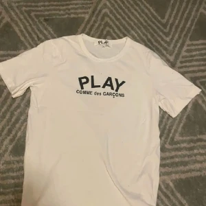 Vit t-shirt från Comme des Garçons - Säljer en vit t-shirt från Comme des Garçons med 'PLAY' tryckt på framsidan och ett stort svart hjärta med ögon på baksidan. Perfekt för en stilren och modern look.