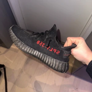 Svarta Adidas Yeezy Boost 350 V2 - Säljer ett par svarta Adidas Yeezy Boost 350 V2 med röd text på sidan. Skorna har en karakteristisk ribbad sula och är tillverkade i ett bekvämt syntetmaterial. Perfekta för dig som vill ha en stilren och modern look. 
