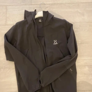 Svart jacka med huva från Haglöfs - Säljer en svart jacka med huva från Haglöfs. Jackan har dragkedja framtill och är långärmad. Perfekt för utomhusaktiviteter med sin stilrena design och funktionalitet.