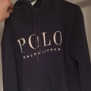Mörkblå hoodie från Ralph Lauren - Säljer en snygg mörkblå hoodie från Ralph Lauren med stor POLO-logga på framsidan och den klassiska polospelaren broderad på ärmen. Tröjan har en justerbar huva med snören och en praktisk magficka. Perfekt för en avslappnad stil.