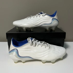  Adidas Copa sense.1  - Snygga vita Adidas Copa fotbollsskor med blå detaljer och snörning. Skorna har en modern design med vågformade linjer och en slitstark sula för optimalt grepp på planen. Perfekta för fotbollsspelare som vill ha både stil och funktion. (Priset kan diskuteras!)