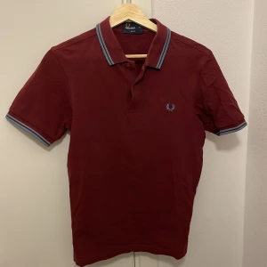 Vinröd pikétröja från Fred Perry - Säljer en stilren vinröd pikétröja från Fred Perry i slim fit. Tröjan har korta ärmar och en klassisk krage med blå och vita ränder. Perfekt för en avslappnad men ändå snygg look.