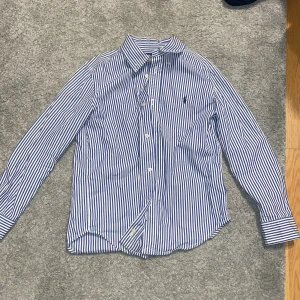 Randig skjorta från Ralph Lauren - Snygg blå- och vitrandig skjorta från Ralph Lauren med klassisk krage och broderad logga på bröstet. Skjortan har långa ärmar och knappar framtill. Perfekt för en stilren look.