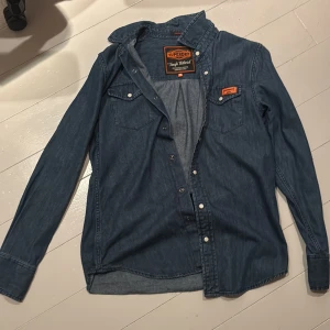 Blå jeansskjorta från Superdry - Säljer en snygg blå jeansskjorta från Superdry med långa ärmar och knappar framtill. Skjortan har två bröstfickor med knappstängning och en cool etikett på bröstet. Perfekt för en avslappnad stil.