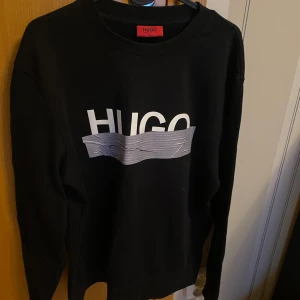 Svart tröja från Hugo Boss | Strl M - Snygg svart tröja från Hugo med ett stort tryck på framsidan. Tröjan har långa ärmar och en klassisk rund halsringning. Perfekt för en stilren look. Den är i gott skick!