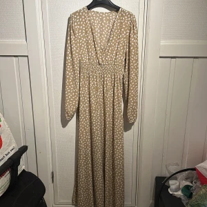Beige långklänning från shein  - Snygg beige långklänning från shein med vita prickar. Klänningen har en v-ringad omlottdesign och långa ärmar med resår vid ärmsluten. Perfekt för en stilren look!