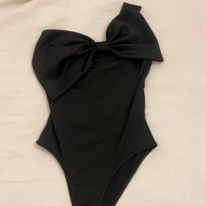 Body med rosett - Oanvänd svart body med stor rosett över ena axeln. Bodyn har knäppe ner till, enkel att ta av och på. Säljer då den ej kommit till användning🤍