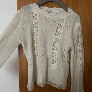 Vit stickad tröja från Zara - Säljer en söt vit stickad tröja från Zara med spetsdetaljer längs ärmarna och framsidan. Tröjan har ett fint mönster och långa armar. Passar bra till både jeans och kjol. 🧶✨