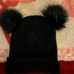 Svart mössa med pälsbollar - Snygg svart mössa med två stora pälsbollar på toppen. Perfekt för att hålla värmen under kalla dagar. Den har en enkel och stilren design som passar till det mesta.