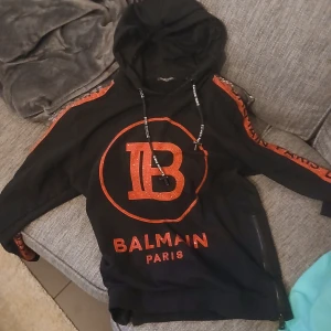 Svart hoodie från Balmain - Säljer en snygg svart hoodie från Balmain med röd glittrig logga och text. Hoodien har dragkedja på sidan och snörning i luvan. Perfekt för en stilren look med en touch av glamour.