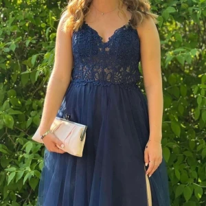 Mörkblå långklänning med spetsdetaljer - Elegant mörkblå balklänning med vacker spets upptill och en luftig kjol. Klänningen har en öppen rygg med snörning och smala axelband. Klänningen har även en slits vid benet.          Perfekt för balen!                                                                   Klänningen är uppsydd och passade perfekt för mig som är 163cm, med klackar med höjden 6,5cm. Klänningen har endast använts 1 gång. Klänningen köptes för 1693kr.