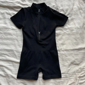 Svart ribbad jumpsuit med dragkedja - Säljer en svart ribbad jumpsuit med korta ärmar och dragkedja framtill. Perfekt för en stilren och bekväm look. Tillverkad i en mix av polyamid och elastan för en stretchig passform. Endast testad. 