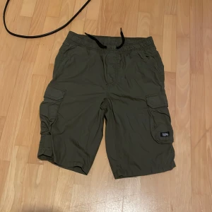 Gröna cargoshorts med snörning - Säljer ett par gröna cargoshorts med elastisk midja och snörning. Shortsen har flera praktiska fickor på sidorna och är tillverkade i 100% bomull, vilket gör dem bekväma och slitstarka.