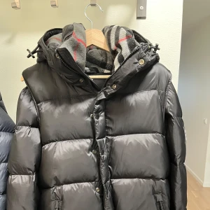 Svart dunjacka från Burberry - Snygg svart dunjacka från Burberry med en klassisk design. Jackan har en avtagbar huva och stängs med dragkedja och knappar. Insidan är fodrad med ett stilrent rutigt mönster. Perfekt för kalla dagar. Aldrig använt helt nyskick. Pris kan diskuteras