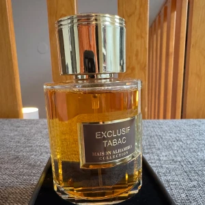 Maison Alhambra Exclusif Tabac  - Exklusiv parfymflaska från Maison Alhambra med doften 'Exclusif Tabac'. Flaskan är elegant designad med en gyllene etikett och ett matchande lock. Parfymen kommer i en stilren kartong med samma gyllene detaljer. Perfekt för den som söker en sofistikerad och unik doftupplevelse.
